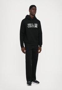 Νέος άνδρας που στέκεται φορώντας μαύρη κουκούλα "Versace Jeans Couture", μαύρα παντελόνια με μπροστινές ραφές και μαύρα αθλητικά παπούτσια με λευκές σόλες.