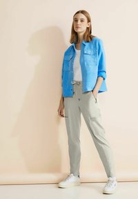 Ljusblå knapp-up skjorta över en vit t-shirt, kombinerad med ljusgrå smalbyxor och vita sneakers. Texturerade material och fickor synliga.