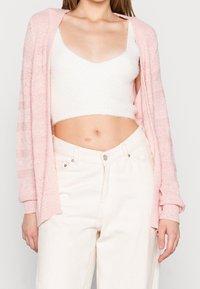 Světle růžový otevřený pletený kardigan přes bílý huňatý crop top. Světlé džíny s vysokým pasem. Hladká textura, volný střih a uvolněný styl.