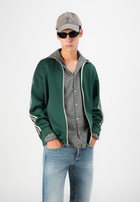 Veste zippée verte avec des accents blancs sur une chemise à carreaux, associée à un jean en denim bleu clair et une casquette de baseball grise.