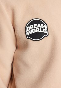 Tissu en polaire beige avec un patch circulaire noir portant l'inscription "DREAM WORLD" en lettres blanches audacieuses.