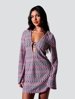 Kvinde med langt mørkt hår iført en langærmet, mønstret pink og lilla minikjole med dyb V-hals og en dekorativ ring ved brystet.