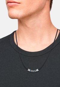 Collana nera intrecciata con accenti argento e motivi geometrici, indossata sopra il colletto di una maglietta scura, che mette in risalto la scollatura.