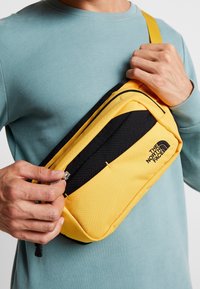 Žlutá a černá kabelka přes rameno s přední kapsou na zip, vyrobená z odolné tkaniny, s logem The North Face a elegantním designem.