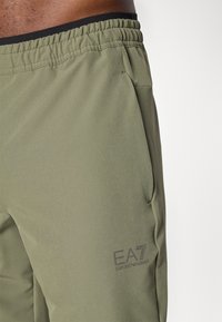 Olive green shorts with an elastic black waistband, side pockets, and a small dark logo "EA7" on the lower left side. Smooth fabric texture.