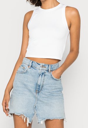 Femme portant un haut court sans manches blanc et une mini-jupe en jean bleu clair effilochée, la main dans la poche.