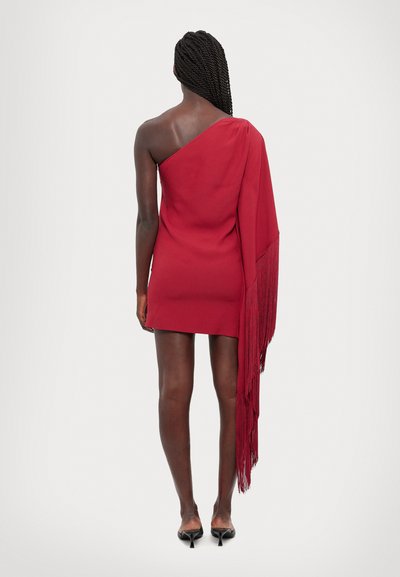 Vestido rojo de un solo hombro, con un largo acento de flecos. Tela suave con un diseño ajustado. Llevado con zapatos de tacón negros.