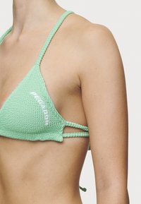 Mintgrønn heklet bikinitopp med trekantkopper, strukturert stoff og flettede stropper. Har hvit logodetalj langs siden.