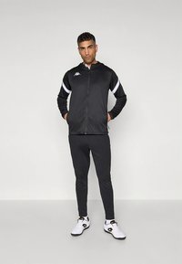 Completo sportivo nero con giacca con cerniera, strisce bianche e logo, abbinata a pantaloni aderenti e scarpe da ginnastica bianche. Tessuto dalla trama liscia.