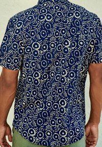 Kratší rukávová košile vyrobená z látky s tmavě modrou základnou a bílými květinovými a listovými vzory. Volný střih s špičatým límcem.