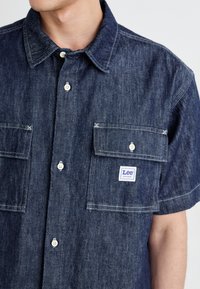Dark denim short-sleeve shirt with two front pockets, white buttons, and a blue "Lee" logo patch on the left pocket. Textured fabric.