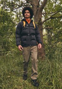 Czarna kurtka puchowa z widocznym szwem, pomarańczowa podszewka, brązowe spodnie cargo i żółte paski plecaka, umieszczona w lesie.