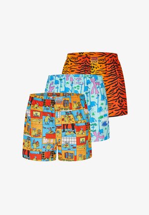 Muskelbepackter Mann mit kurzen Zöpfen, der hellblaue Boxershorts mit bunten Dinosauriermustern trägt, steht vor einem schlichten hellen Hintergrund.
