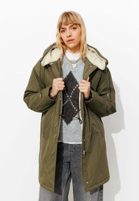 Parka verte avec capuche bordée de fourrure, fermeture éclair et taille ajustable par cordon, portée sur un pull gris à motif de diamants et un jean ample.