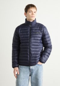 Navy puffer jacket with a high collar, horizontal quilted design, front zipper, and small logo patch on the left chest.