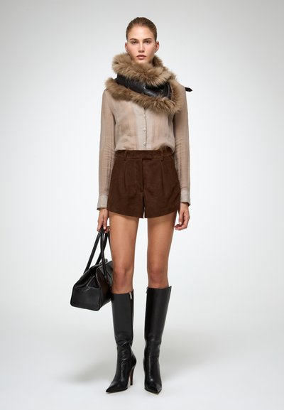 Donna che indossa una blusa trasparente, pantaloni corti in suede marrone, sciarpa con bordo in pelliccia, stivali neri fino al ginocchio e tiene in mano una borsa in pelle nera.