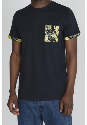 Uomo che indossa una T-shirt nera a maniche corte con stampa di foglie tropicali sulla tasca sul petto e sui polsini delle maniche, abbinata a jeans blu.