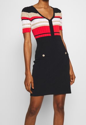 Kobieta ubrana w sukienkę z krótkim rękawem z głębokim dekoltem w kształcie litery V, z czerwono-biało-złoto-czarnym paskowanym topem oraz gładką czarną spódnicą z detalami w postaci złotych guzików.