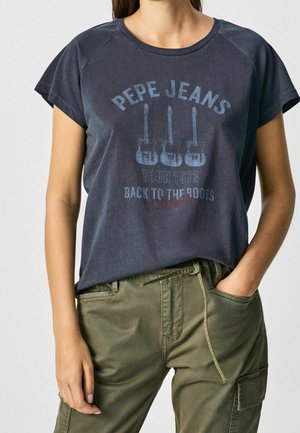 Kobieta w ciemnoniebieskim T-shircie Pepe Jeans z trzema gitarami i napisem „Tour 1973 Back to the Roots London” w połączeniu z oliwkowozielonymi spodniami typu cargo.