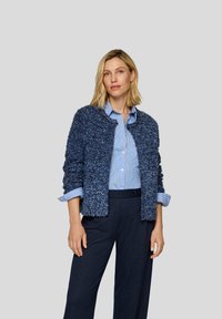 Navy strukturierte Strickjacke mit einem flauschigen Finish über einem hellblau gestreiften Hemd, kombiniert mit dunkel gestreiften Hosen. Lässiges und geschichtetes Outfit.