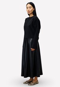 Zwarte top met lange mouwen en een cropped ontwerp, gecombineerd met een los vallende zwarte rok van nepleer en metalen platte schoenen met een decoratieve strik.