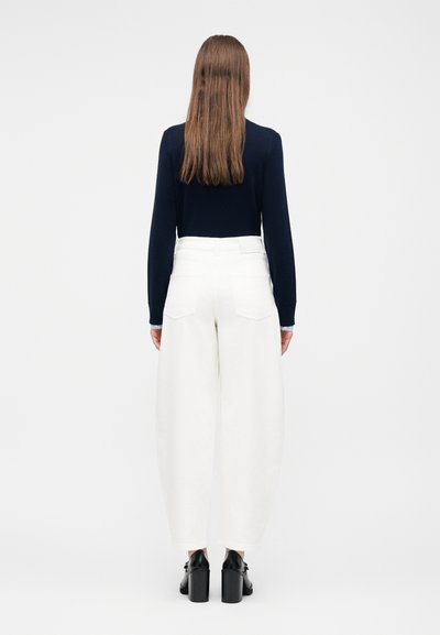 Γυναίκα με μακριά καστανά μαλλιά, γυρισμένη προς τα πίσω, φοράει μαύρο μακρυμάνικο τοπ, λευκό παντελόνι φαρδύ στο κάτω μέρος και μαύρα παπούτσια με τακούνι.