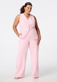 Lichtroze getailleerde vest en wijde broek gemaakt van een gladde stof. Vest heeft knopen en zakken. Model draagt accessorized sieraden.