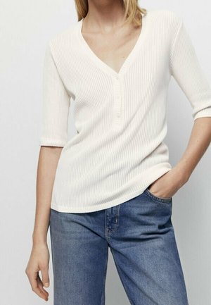 White, ribbed top with a V-neck and three-quarter sleeves, featuring a button placket and a relaxed fit, paired with blue jeans.