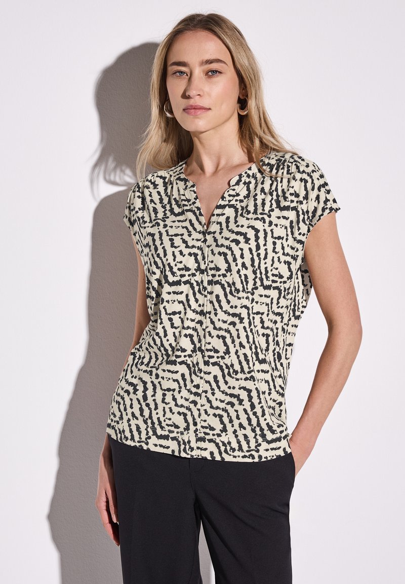 Blusa a maniche corte color crema con un motivo astratto nero. Presenta un design senza colletto e un tessuto morbido e leggero.