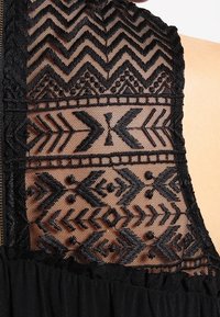 Szczegół czarnej sukienki z przezroczystym panelem z koronki z geometrycznym haftem, obejmującym szewrony i kropki, na obszarze ramienia.