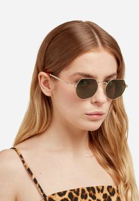 Okrugle sunčane naočale s tamnim staklima, tankim srebrnim metalnim okvirom i narandžasto naglašenim krakovima. Naočale nosi model s dugom, ravnom kosom.