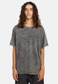 Camiseta de manga corta y cuello redondo en denim gris oscuro con textura desgastada. Presenta costuras en contraste y un acento de parche de cuero en el dobladillo.