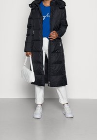 Sort puffervinterjakke med hætte, der har en frontlynlås, sidelommer og et langt, quiltet design. Parret med hvide bukser og sneakers.
