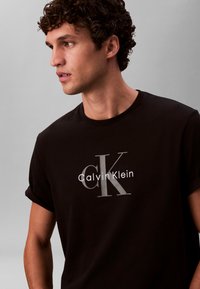 Crna pamučna majica kratkih rukava s uvrnutim rubovima i središnjim logom s natpisom "Calvin Klein" i velikim "K" u kontrastnoj sivoj i bijeloj boji.