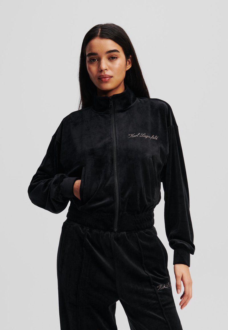 Chaqueta de chándal de terciopelo negro con un alto cuello, cierre frontal de cremallera y logotipo bordado. Presenta un ajuste relajado y puños elásticos.