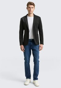 Czarna marynarka z klapami z wycięciem, dwie przednie kieszenie, noszona na białej koszulce i niebieskich dżinsach, zestawiona z białymi trampkami.