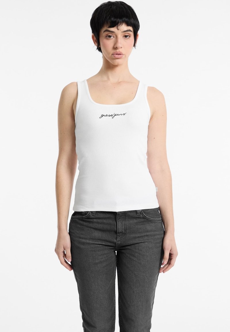 White ribbed tank top featuring a rounded neckline and a black "guessjeans" logo on the front, paired with dark gray jeans.