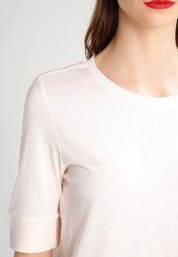 Jasno beżowa koszulka z krótkim rękawem wykonana z miękkiego materiału, z okrągłym dekoltem i podwiniętymi mankietami. Gładka tekstura z minimalnymi detalami wzorniczymi.