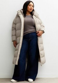 Mujer vistiendo un abrigo acolchado largo beige sobre un suéter de canalé gris, jeans oscuros de pierna ancha y zapatillas blancas, de pie contra una pared blanca.