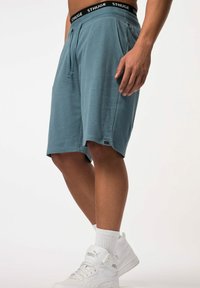 Blauwe sportshorts van zachte stof, met een trekkoord in de taille, zijzakken en een logo-opdruk op de tailleband. Draagt witte sneakers.