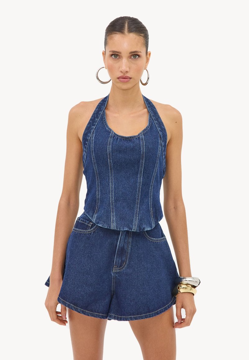 Femme aux cheveux plaqués en arrière portant de grandes boucles d’oreilles en créoles, un haut sans manches en jean à col halter, un short en jean assorti et des bracelets au poignet droit.