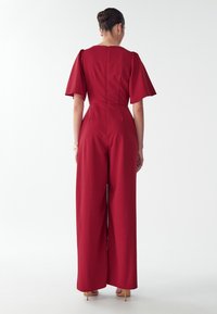 Rode jumpsuit met korte pofmouwen, wijde pijpen, gestructureerd materiaal en een ritssluiting aan de achterkant. Draagt met naakthakschoenen.