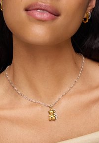 Srebrny naszyjnik z łańcuszka z zawieszką w kształcie złotego misia świątecznego o błyszczącej teksturze, szczegółową twarzą i małym srebrnym akcentem za misiem.