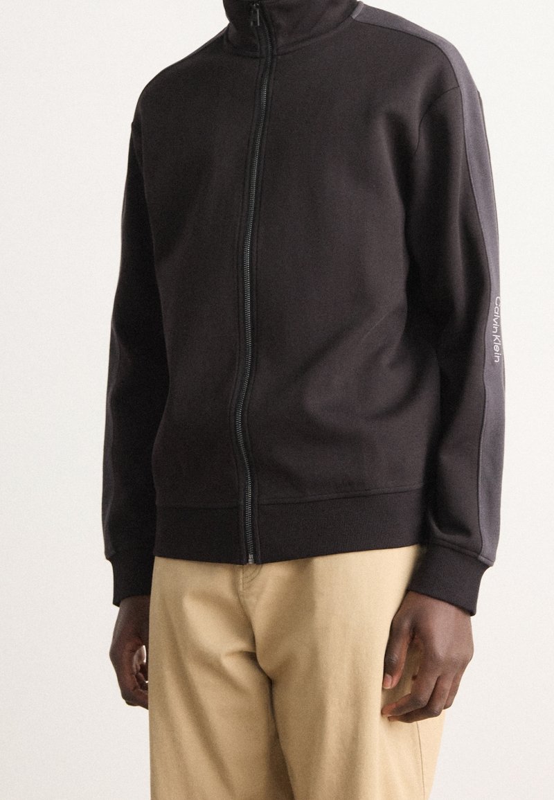 Personne portant une veste noire zippée avec "Calvin Klein" sur la manche et un pantalon beige, debout devant un fond uni.