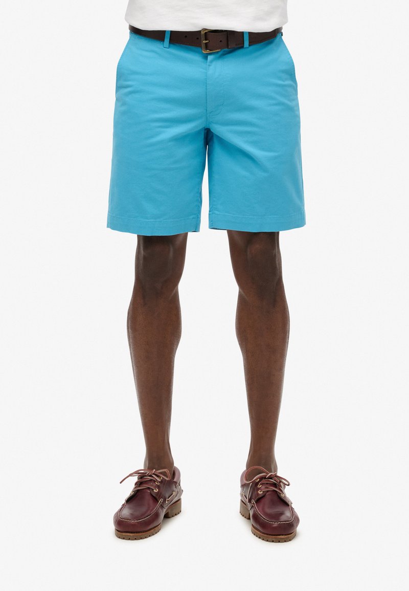 Shorts turquoise avec une texture lisse, présentant une coupe droite et une ceinture en cuir marron. Associés à des chaussures bateau bordeaux foncé.