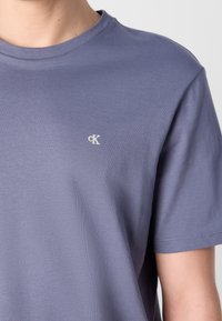 Gros plan d'une personne portant un t-shirt violet uni à manches courtes avec un petit logo blanc "cK" sur le côté gauche de la poitrine.