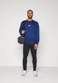Mörkblå sweatshirt med vitt logo, parat med svarta byxor med vita sidostrand. Modellen håller en liten svart väska, bär vita sneakers.