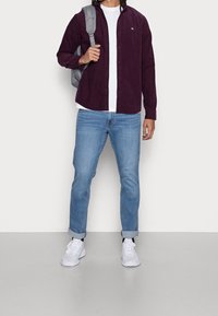 Camicia in corduroy bordeaux, t-shirt bianca, jeans azzurri chiari e sneakers bianche. Zaino grigio appeso alla spalla destra. Design semplice.