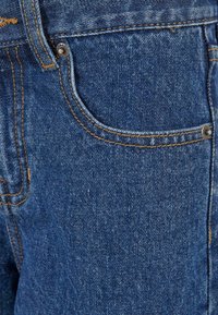 Prim-plan cu buzunarul frontal al unei perechi de blugi albaștri din denim, cu cusături galbene și nituri metalice.