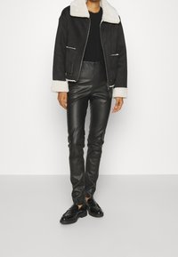 En person bär en svart fårskinnsjacka med vit trim, en svart tröja och tighta svarta läderbyxor, tillsammans med svarta skor.